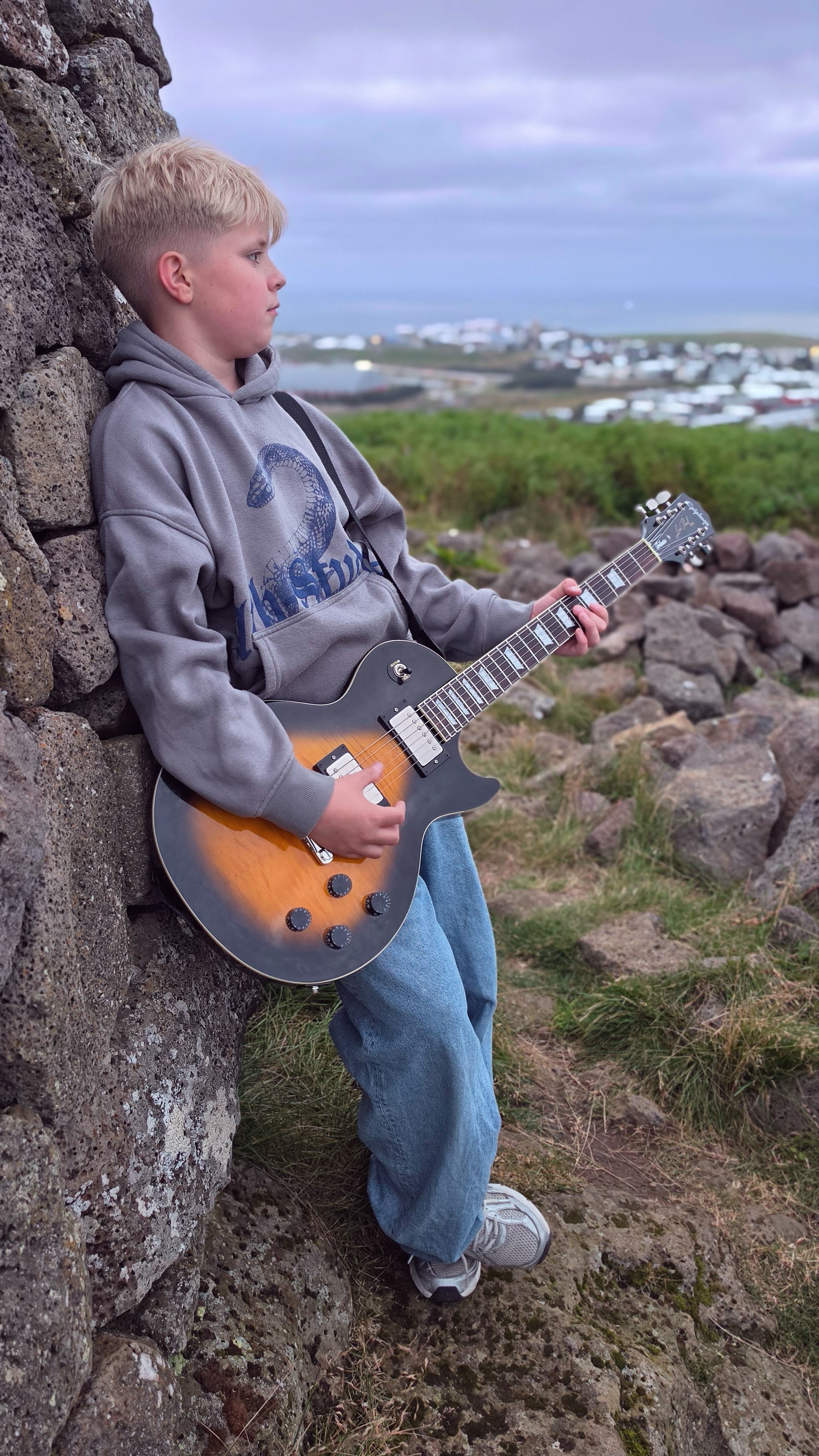 Matthias Thor playing an electric guitar beside a rocky outcrop, with a settlement visible in the distance beneath a cloudy sky.