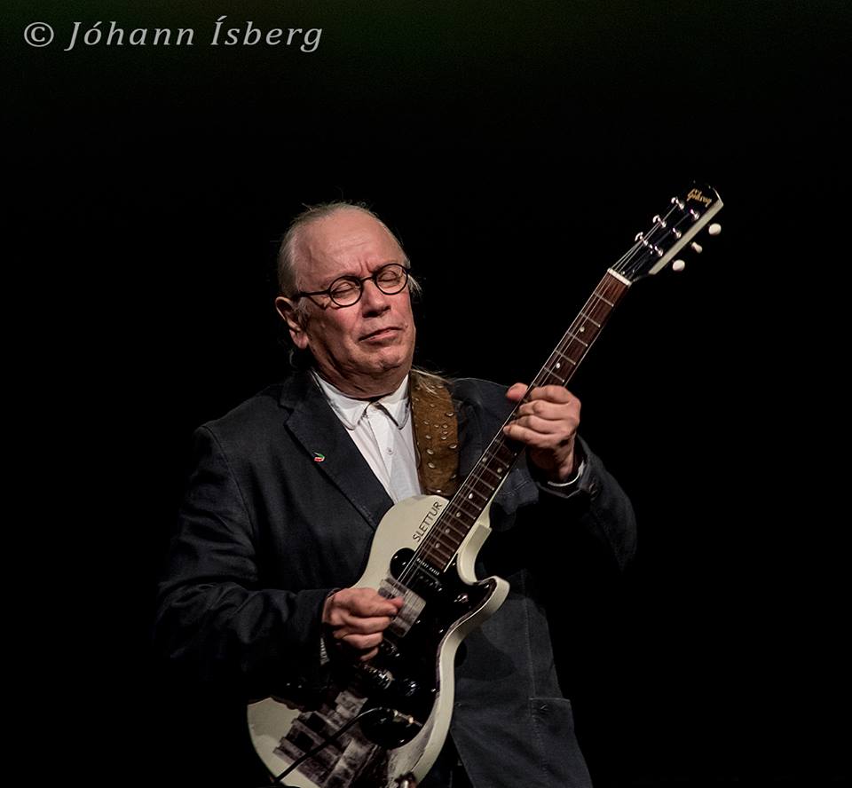 An older man in a dark suit plays an electric guitar on a dimly lit stage.