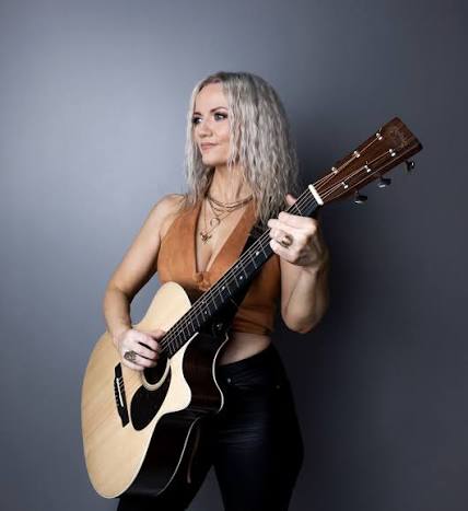 Brynhildur Oddsdóttir holds an acoustic guitar and looks to the side against a dark background.